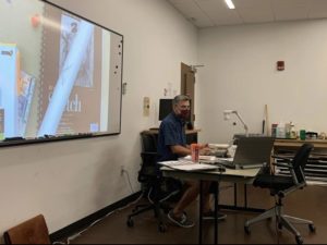 A teacher in his classroom teaching graphic design by using a projector and having the image on a screen behind him