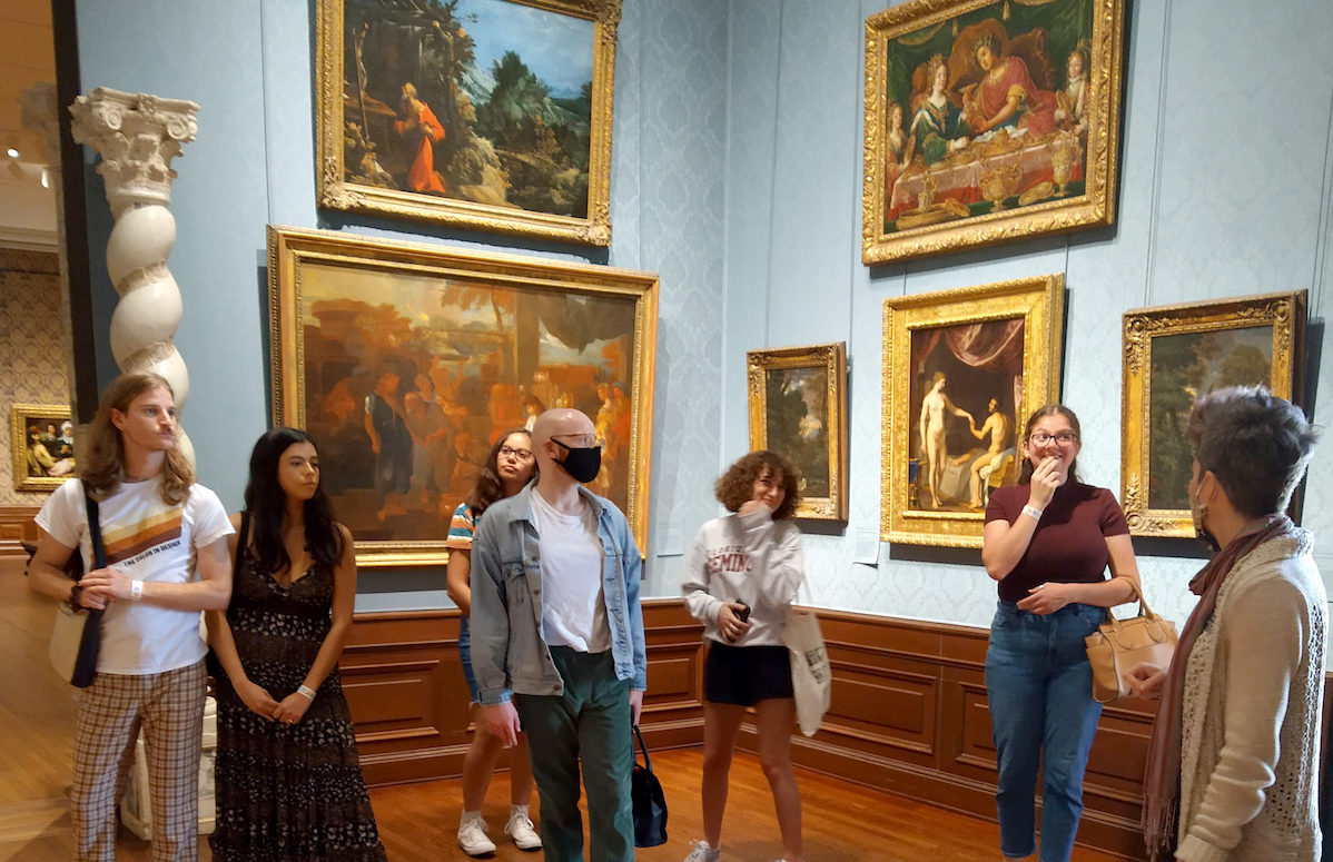 College students and their professor stand in a museum gallery room with blue walls covered in paintings. 