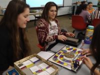 Students sit at a table. They are playing a colorful board game.
