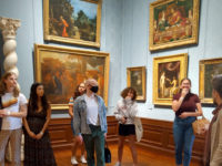 College students and their professor stand in a museum gallery room with blue walls covered in paintings.